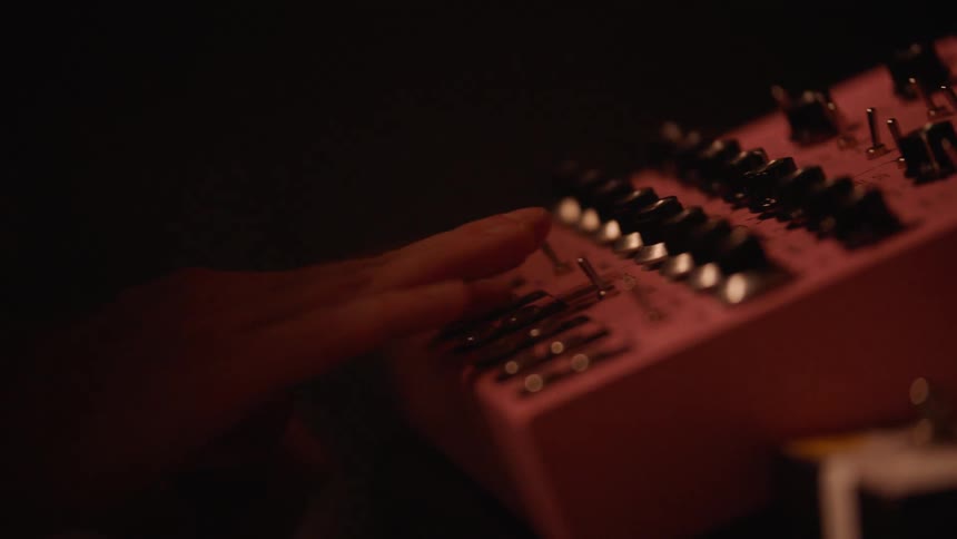 Hands adjust knobs and switches on a synthesizer under intense red lighting