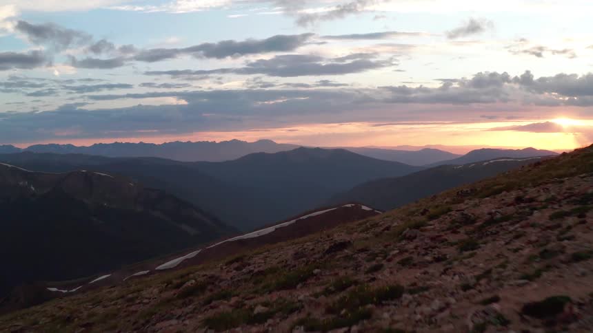 Three people running on top of a mountain in a mountain range with the sun shining in the sky in the morning