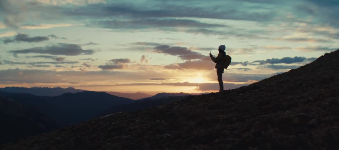 Person with a backpack and beanie watches the sunset from the top of a mountain