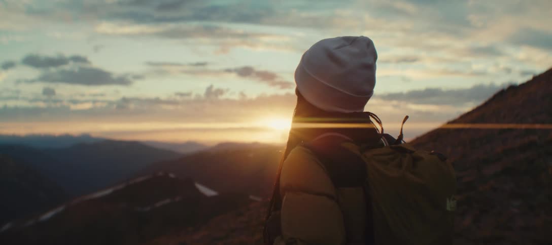 Aq woman wearing a white bonnet is standing looking at a landscape with the sun shining brightly on the horizon
