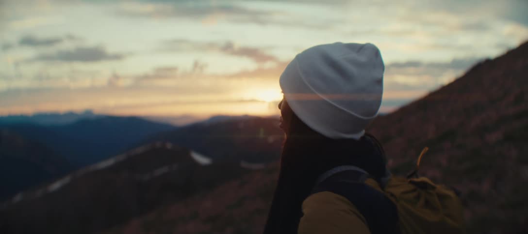 A person with a beanie seen from a side angle standing on a mountain peak and looking out at the beautiful sunset over the distant mountain range