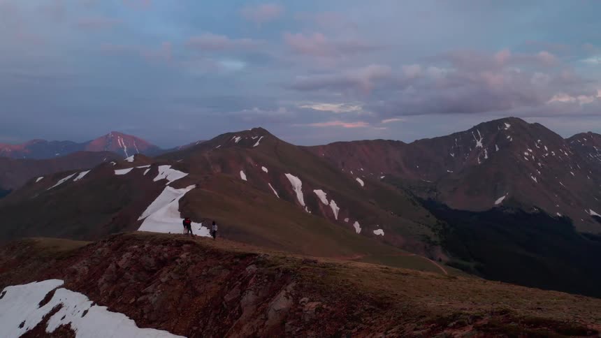 Two people walking on top of a mountain in a mountain range with some snow on some mountains