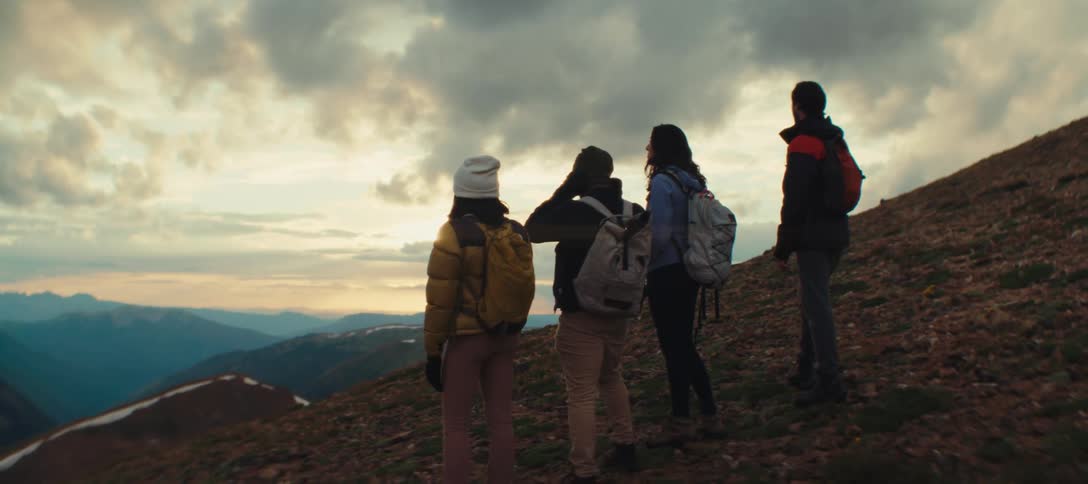 Four people with backpacks watch the mountainous landscape at sunset facing the horizon lit by the setting sun