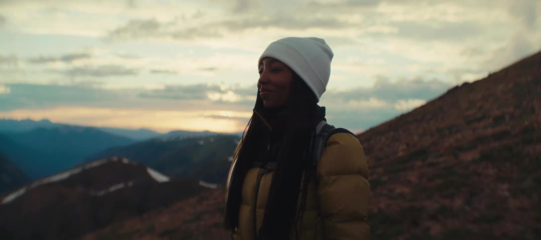 A woman standing wearing a yellow jacket and a white beanie outdoors near a mountain range
