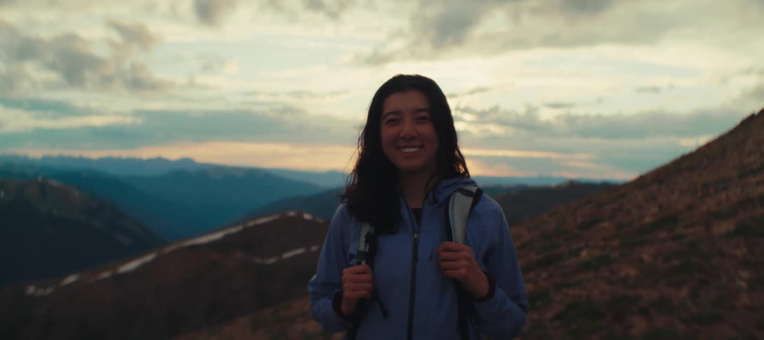 Woman with a backpack smiles while posing on a mountain trail at sunset