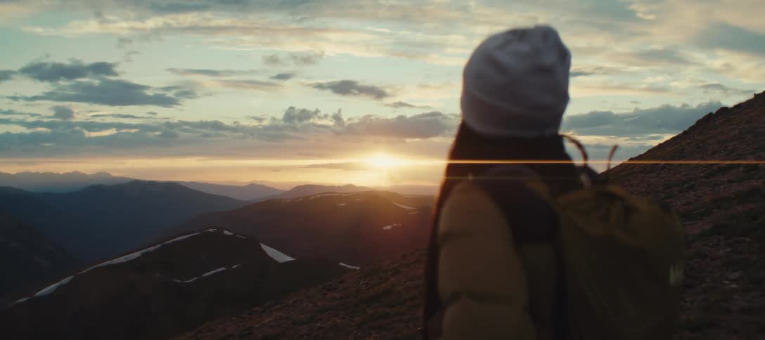 Silhouette of a person with a backpack and beanie looks at mountains at sunset