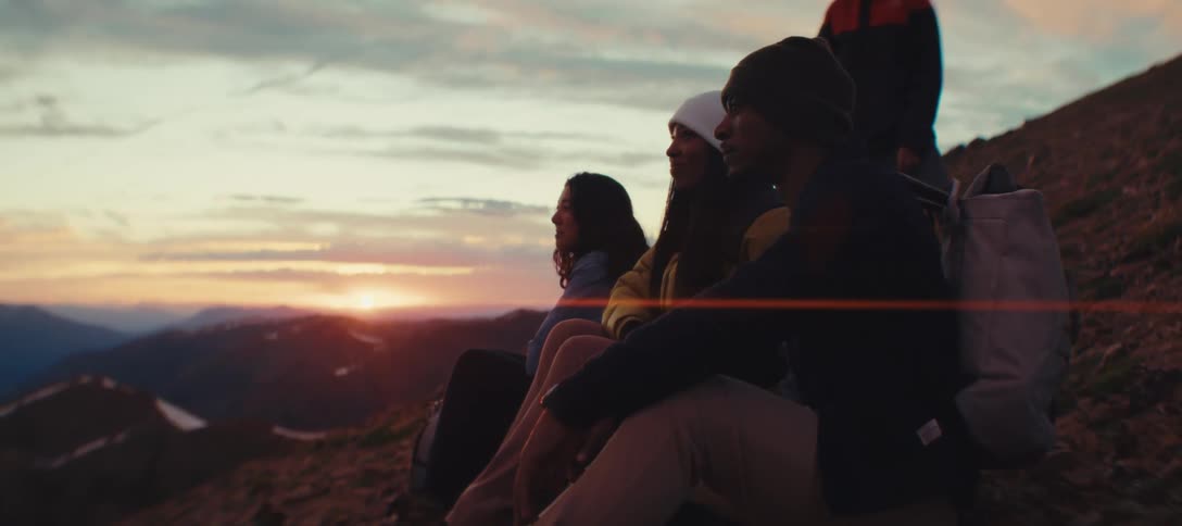 A group of people sit on a rocky hillside facing left and watching a sunset over a distant mountain range while a fourth person stands behind them with a backpack on their back