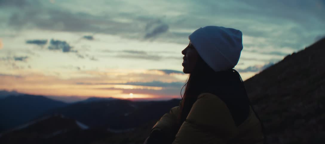 A woman wearing a white beanie is standing outdoors looking at the horizon with the sun shining in the morning sky