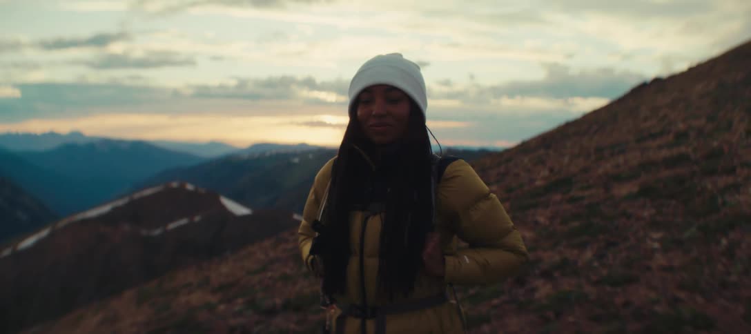 A young black woman stands outdoors on a mountain slope at sunset