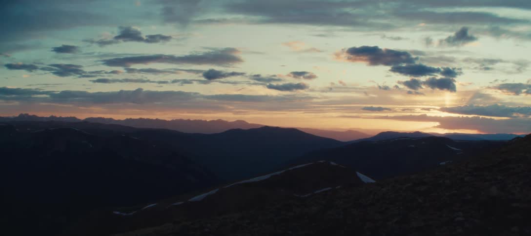 A landscape of a mountain range that is dark in the morning with the sun shining on the horizon in a sky full of white clouds