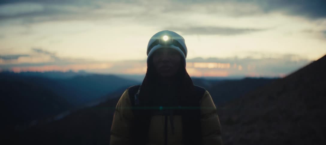 Person in a yellow jacket and white beanie wears a lit headlamp at dusk in the mountains