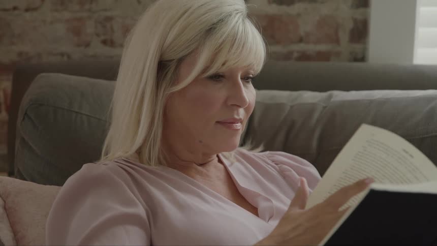 Close up of a blonde woman reading a book while sitting on a couch in a living room during daytime