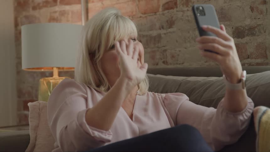 A woman smiles and waves while on a video call with a mobile phone as she sits on a couch in a living room