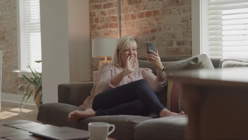 A woman with blonde hair sits on a gray sofa in a brightly lit room with a brick wall and a large window waving and smiling at a cell phone she holds up to her face