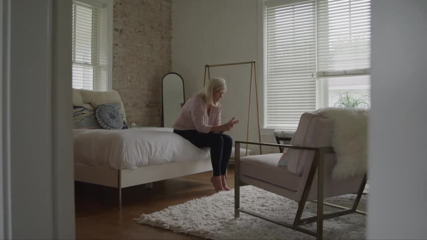 A woman with blonde hair sits on the edge of a bed with white sheets and a blue patterned pillow rubbing her hands while a large mirror a clothing rack and a chair with a white fur blanket are visible in the bright room behind