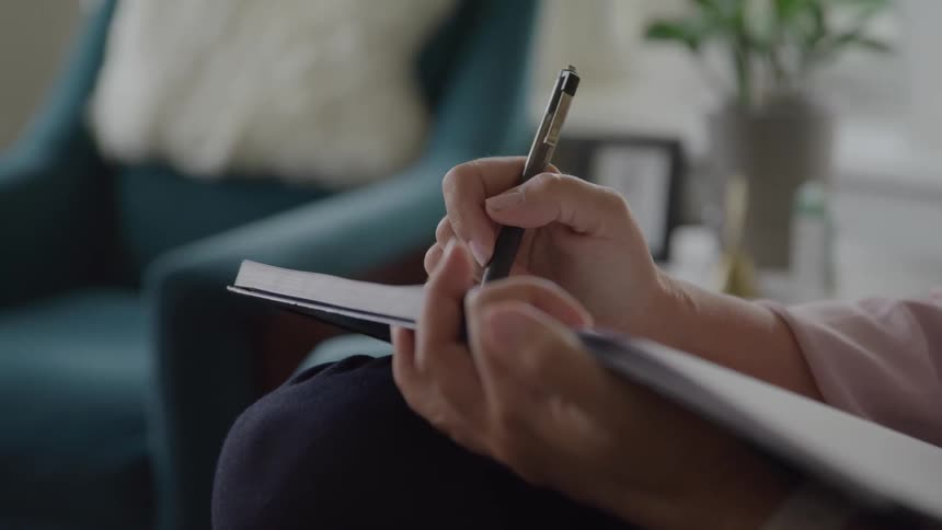 A persons hand using a pen to write on a paper book that is resting on their legs