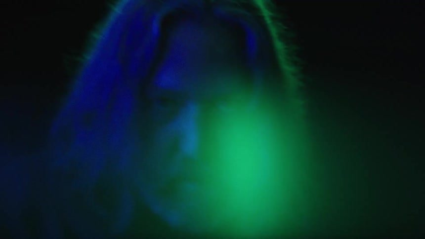 A long haired man stares intensely in blue green light