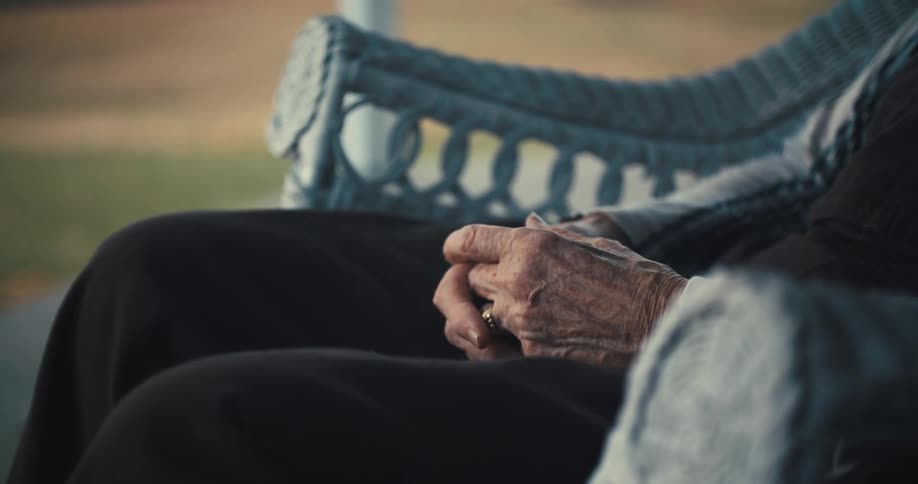An elderly person sits outdoors with hands resting gently together