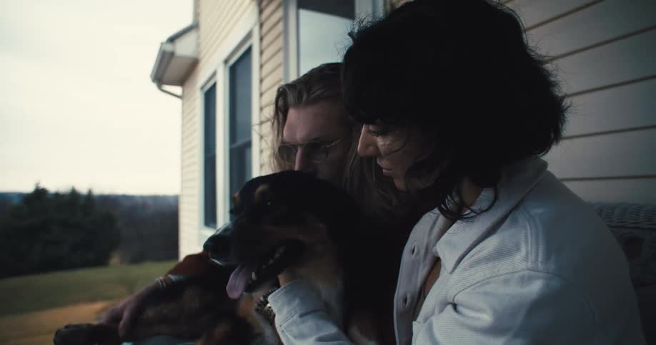 The video shows two people seated on a porch with a dog between them they are petting and interacting with the dog which has its mouth open and tongue hanging out appearing to be panting or smiling