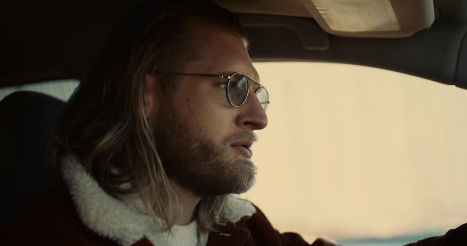 A man with long blonde hair and a beard is sitting in the drivers seat of a car he is wearing a brown jacket with a white collar and goldrimmed sunglasses