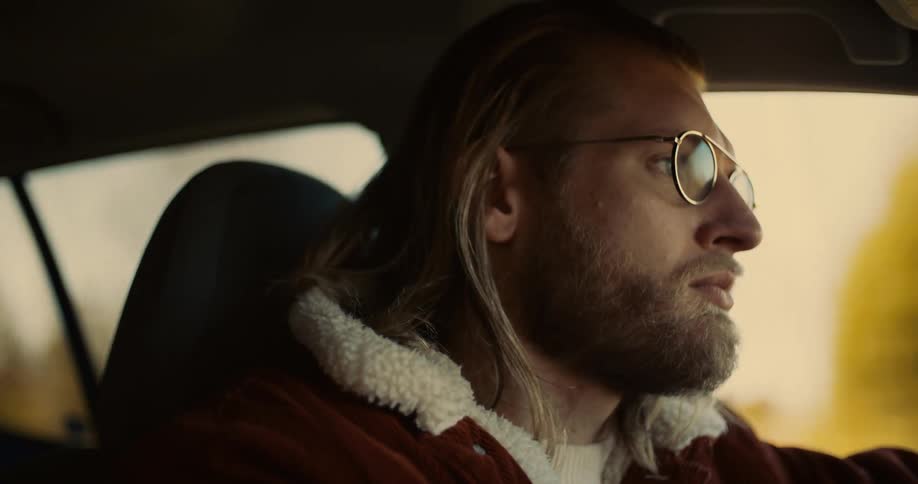 The video shows a man with long lightcolored hair and glasses wearing a brown jacket with a white fur collar in the drivers seat of a car he is looking out the window and the camera captures him from the passengers side
