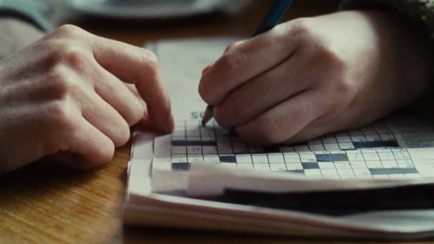 A persons hands are visible on a wooden table one hand is holding a pencil the pencil is used to write on a paper with a partiallyfilled crossword puzzle