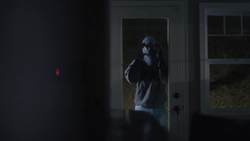 A robber wearing a balaclava peeks into an urban suburban house at night through a door with a large glass pane
