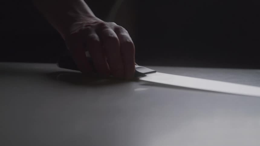 A dark shot of a hand picking up a knife with a black handle from a surface a single beam of light illuminates the knifes blade creating a strong contrast