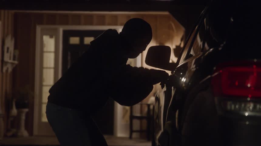 A shadowy figure in a hoodie is seen from the side trying to open the door of a parked car the scene is dark with the illuminated doorway of a house visible in the background