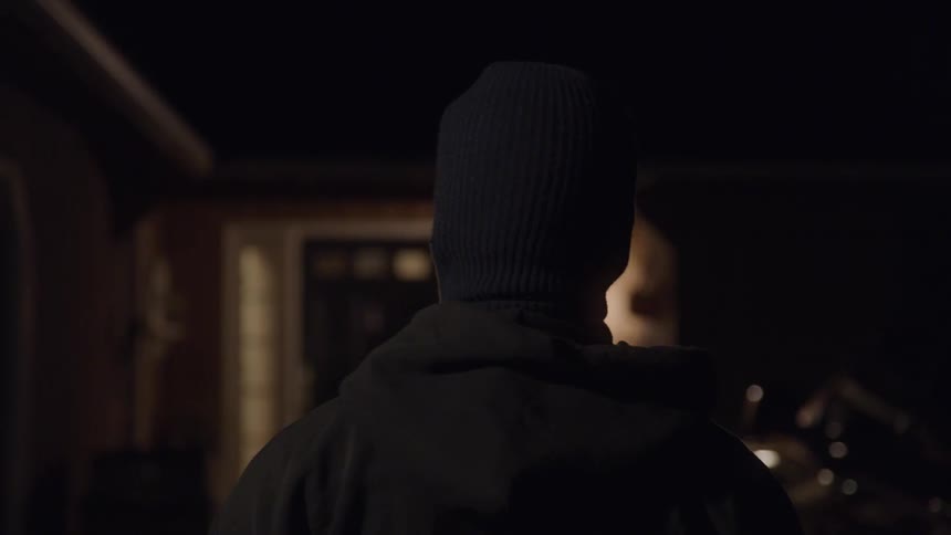A person wearing a jacket and a beanie hat stands in front of a residential home at night with the buildings lights reflecting on the surroundings
