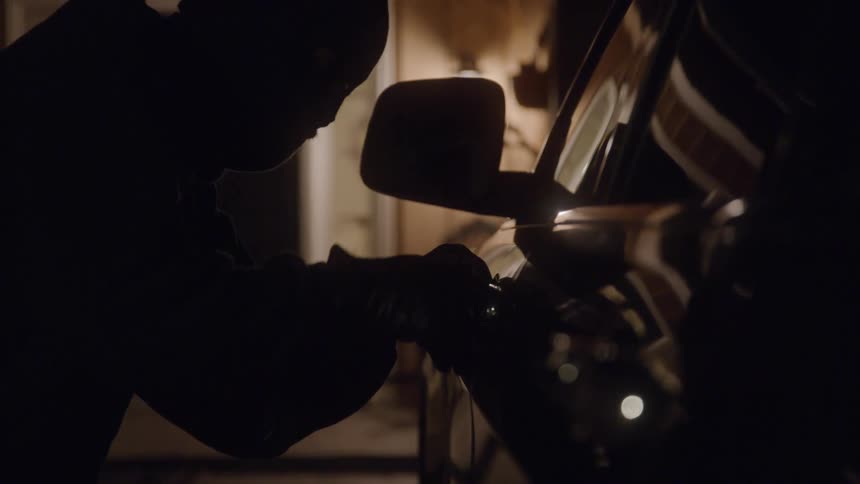 A dark shot of a person trying to open a car door at night the persons silhouetted hands and head are visible as the cars interior lights turn on briefly illuminating them