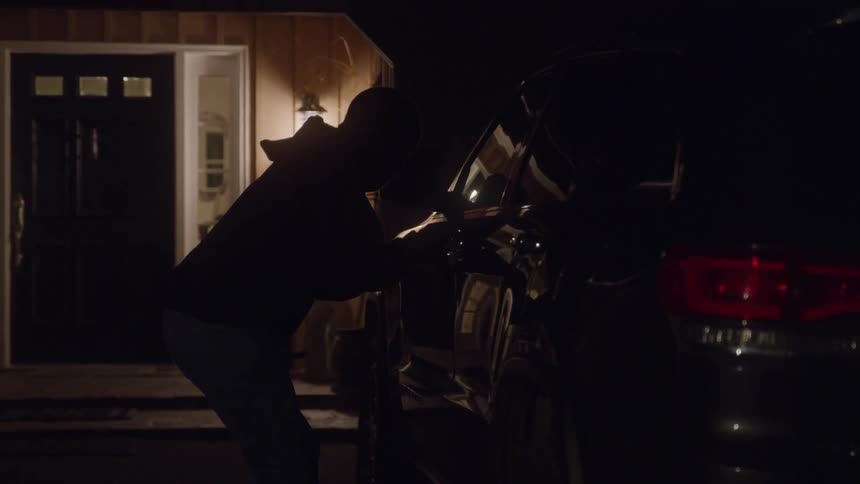 A shadowy thief wearing a hoodie and pants approaches and leans into the drivers side of a dark vehicle with its door open and red tail lights glowing stealing the car while parked in front of a house with a lit porch