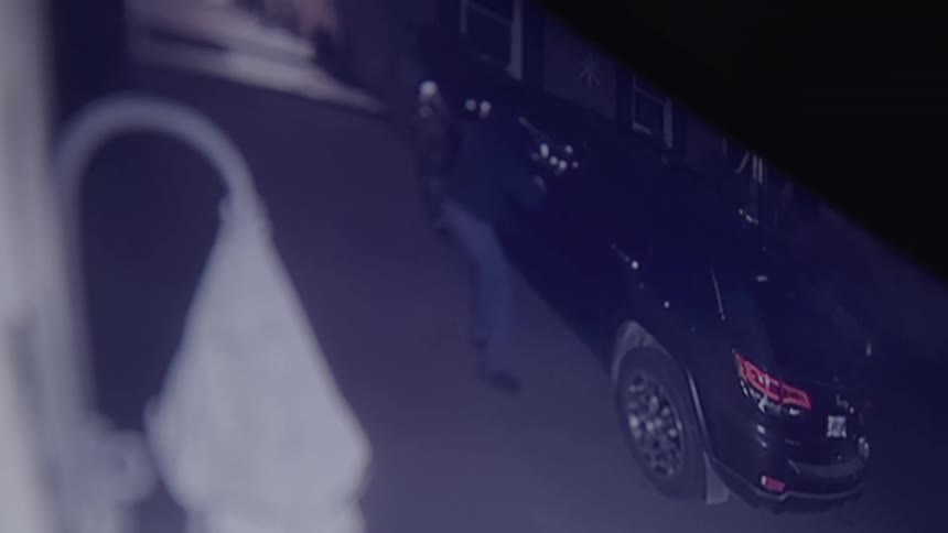 A burglar wearing a hoodie creeps up to a car and breaks in
