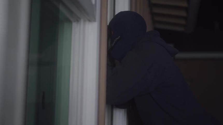 A person wearing a black ski mask and hoodie is seen peeking through a window