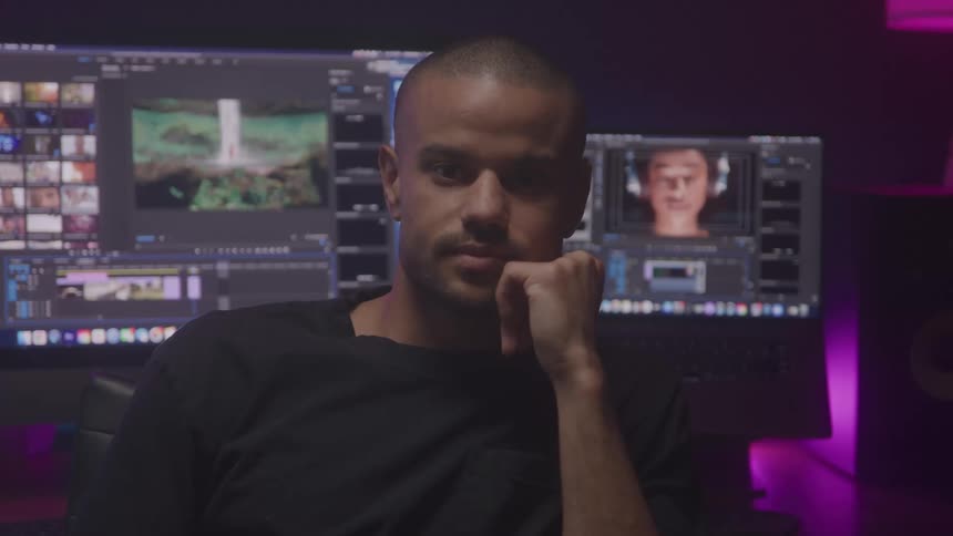 Man sits with his back to his computers while in an office lit by purple lighting