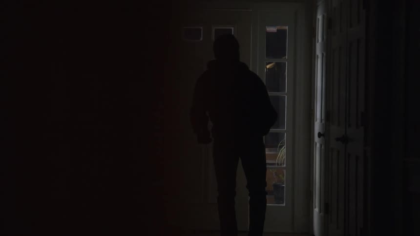 A masked intruder walks through the front door of a house while carrying a knife