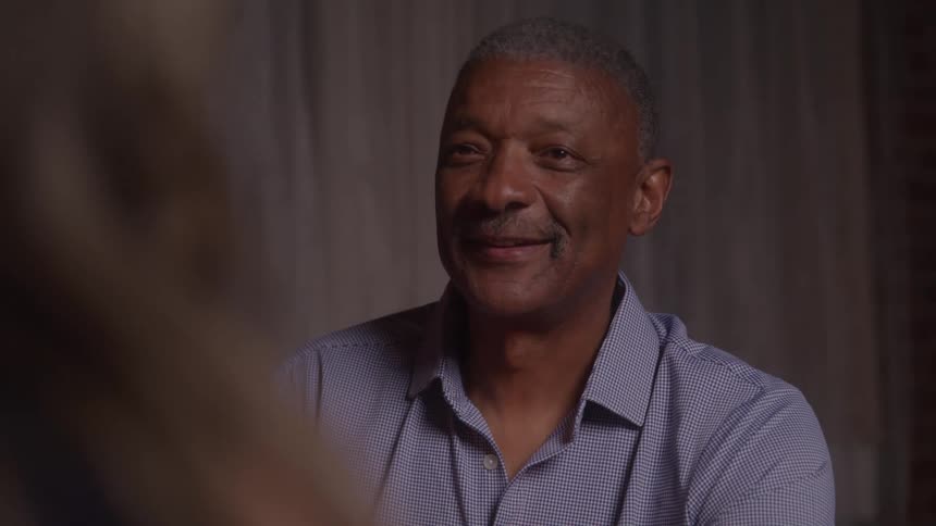 A smiling older black man with a saltandpepper haircut is seen from the chest up he is wearing a blue checkered shirt and looking off to the side seemingly engaged in a conversation with someone just out of frame