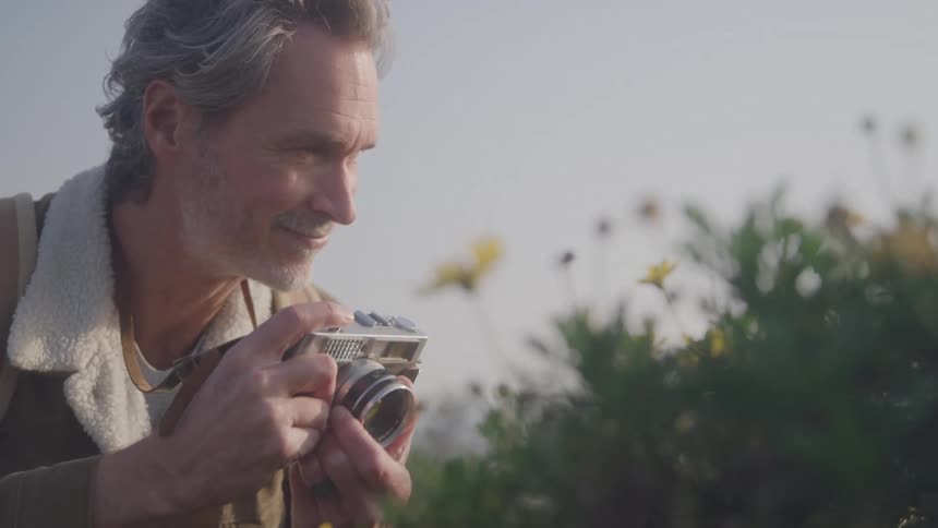 Man wearing a jacket takes a picture of a flowering bush during the daytime