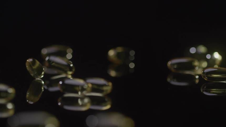 Golden translucent softgel capsules fall and bounce in dramatic slow motion onto a black reflective surface that clearly mirrors their movement