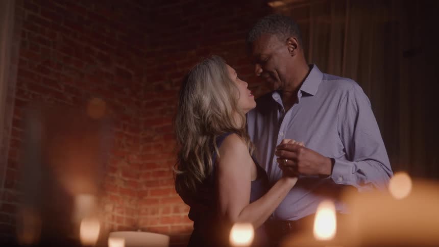 A mature couple dances slowly holding hands and looking into each others eyes surrounded by glowing candles and a brick wall