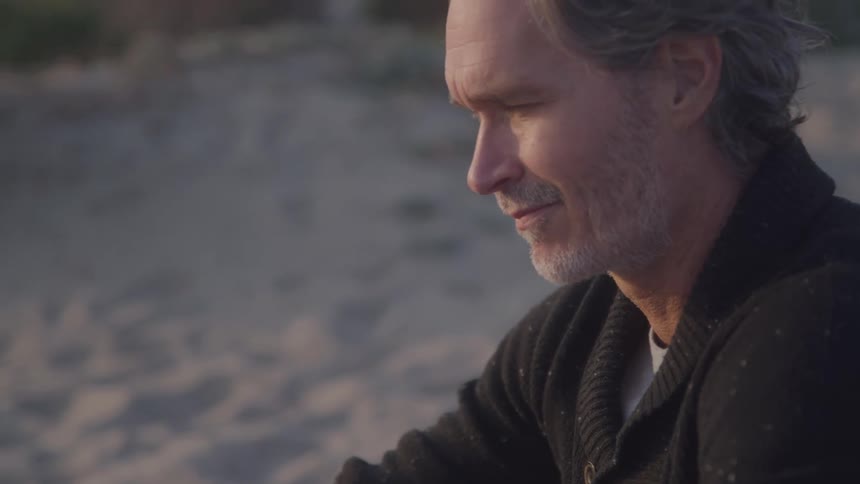 A mature man with graying hair and a beard is shown in a closeup side profile view he is smiling faintly and looking offcamera with a softfocus beach background behind him