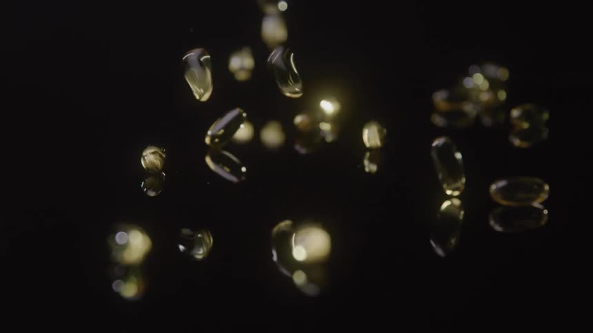 Numerous yellow oblong gelatin capsules fall and bounce on a dark reflective surface creating bright glints of light before coming to a rest