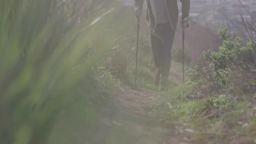 A hiker is walking down a grassy path on a hillside using two hiking poles the shot starts as a wide shot and zooms in focusing on the persons feet as they continue walking