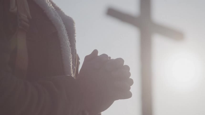 A person is seen in a profile shot with their hands clasped together in prayer a blurry cross is visible in the background against a bright hazy sky