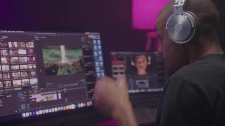 A person with a shaved head is shown from the side as they put on a pair of silver headphones they are sitting in front of two computer monitors seemingly preparing to edit video