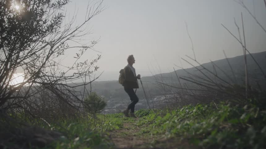 A silhouetted man with a backpack and trekking poles hikes along a narrow dirt trail on a hillside backlit by the bright low sun of a hazy sunrise or sunset