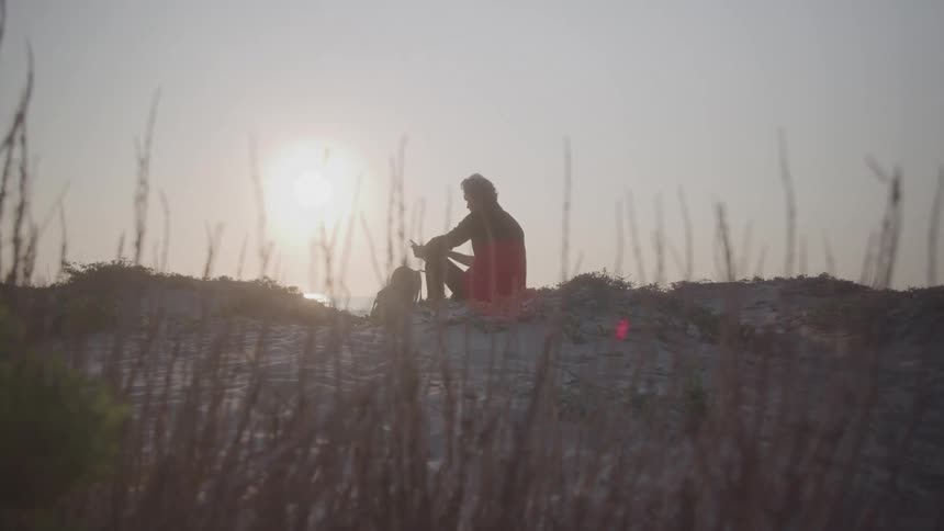 A silhouetted man with a backpack sits on top of a sandy hill and reads something while backlit by the bright hazy glare of the low sun