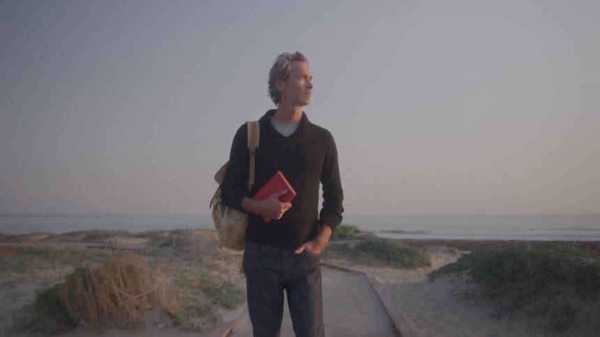 The man is walking on the beach holding a book