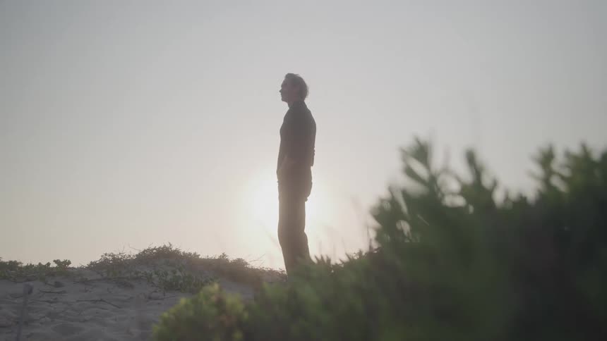 A side view of a man in silhouette stands on a sandy hill and looks left with the bright sun behind him and some green bushes in the foreground