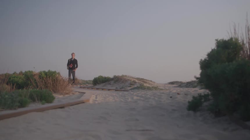 A wide shot of a man standing alone on a boardwalk surrounded by sandy dunes and lowlying bushes looking out at the horizon